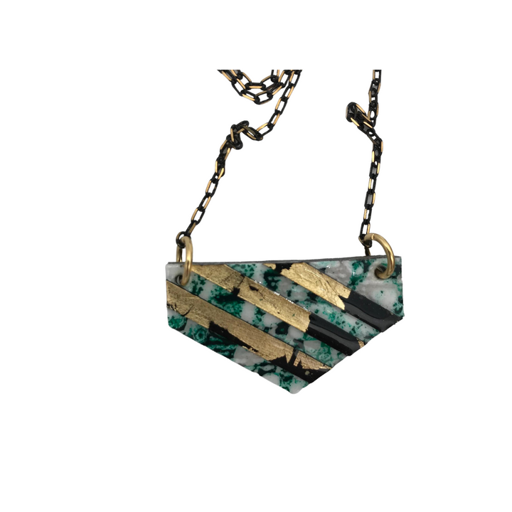 The Rothlú Ralston Batik Textile Necklace in Dark Green/Black/Gold features a stylish pendant shaped like an inverted trapezoid, showcasing a marbled pattern of green and white with diagonal stripes of gold and black. Enhanced with a high gloss eco resin finish, the black chain has a subtle pattern and connects to the pendant with gold rings.