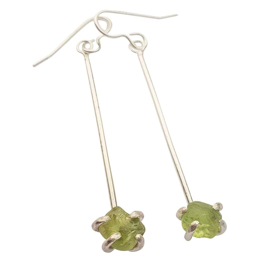 A pair of Raw Peridot Drop Earrings by Caroline Stokesberry-lee with long, thin sterling silver rods ending in raw Peridot gemstone accents held by silver prongs. The earrings have hook-style ear wires for wearing.