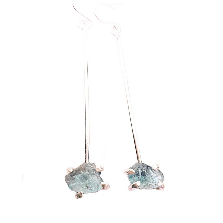 Caroline Stokesberry-lee's Raw Aquamarine Drop Earrings: A pair of long, sterling silver drop earrings featuring rough, uncut aquamarine gemstones at the bottom. The stones are held in place by prong settings attached to delicate ear wires, creating a simple yet elegant design against a white background.