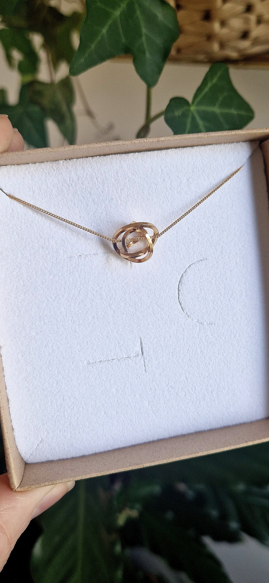The Caroline Stokesberry-lee Knot Pendant Gold necklace, handcrafted and plated in 18ct yellow gold, is shown in a brown box with white lining. Green leaves in the background highlight this elegant jewelry piece.