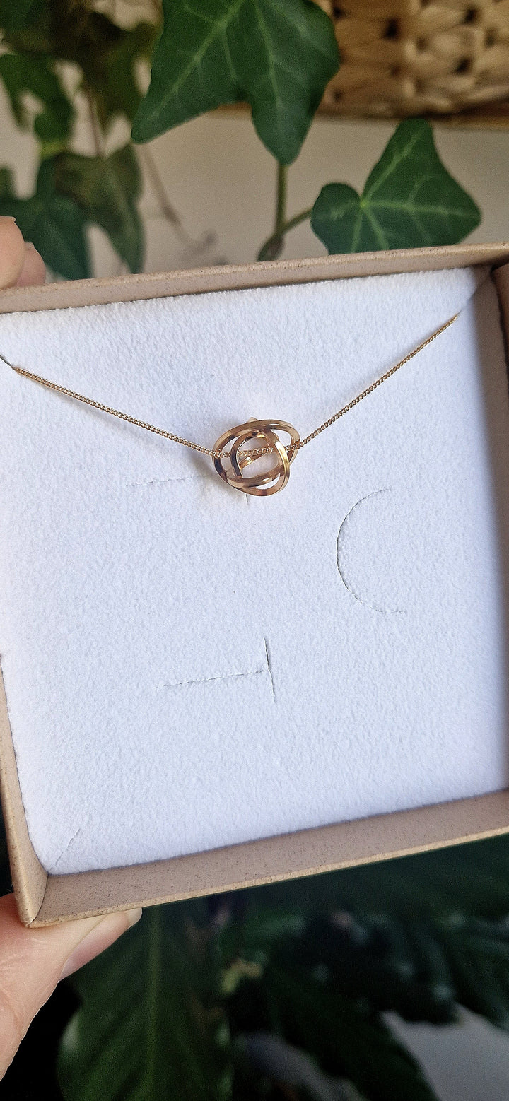 The Caroline Stokesberry-lee Knot Pendant Gold necklace, handcrafted and plated in 18ct yellow gold, is shown in a brown box with white lining. Green leaves in the background highlight this elegant jewelry piece.