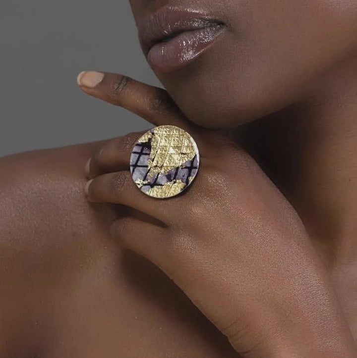 A person’s hand, near their chin, wears the Rothlú Ró Cocktail Ring in Gold/Black—a bold, adjustable statement piece with a geometric design—against a plain, neutral background.