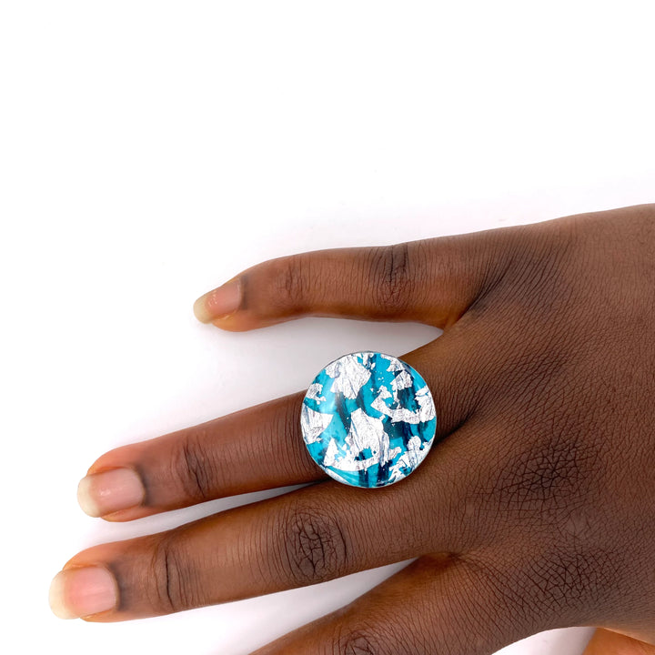 A hand with dark skin wears the Rothlú Ró Beag Dress Ring in Silver/Sea Blue, featuring a large, round sgraffito batik design with blue and silver marbling, shown against a plain white background.