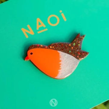 The Robin Pin Brooch by Naoi is a colorful enamel pin shaped like a robin, with an orange breast, sparkling brown wings, and a glossy resin finish. It’s displayed on a teal card with "Naoi" printed above it.