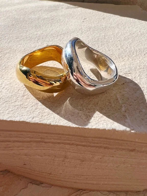 A close-up of two polished, chunky gold and silver Rocco Rings by Shyla resting on a textured stone surface. They both have smooth, curvy, and slightly irregular shapes, casting soft shadows.