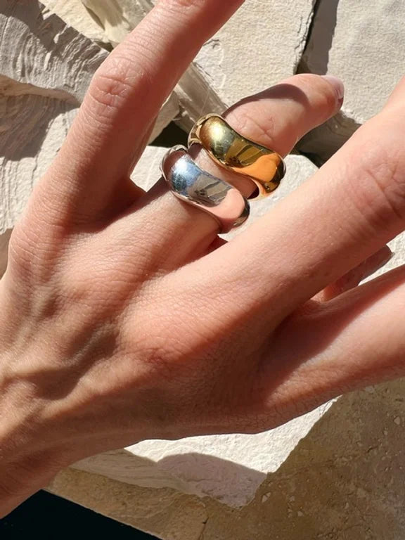 Close-up of a hand with two chunky, stackable Rocco Rings by Shyla, one gold and one silver, worn on the middle and ring fingers. The hand is resting on a textured, light-colored surface, with sunlight casting shadows and highlighting the metallic sheen of the rings.