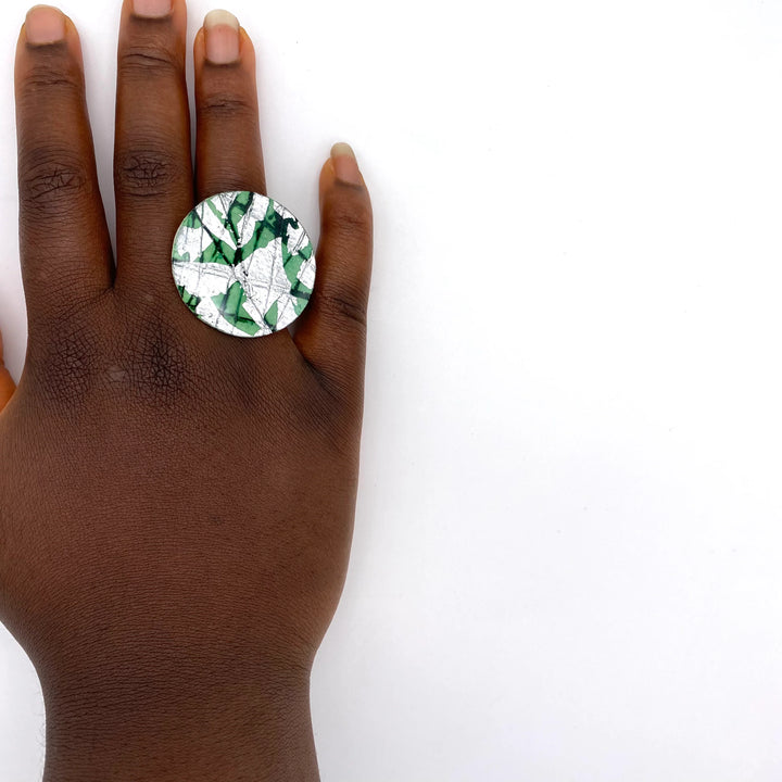 A hand with medium-dark skin tone wears the Rothlú Ró Cocktail Ring in Silver/Jade—a large, hypoallergenic silver-plated ring featuring a green and white abstract pattern—shown against a plain white background.
