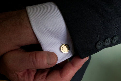 A close-up image of a person's hand adjusting the cuff of a white dress shirt. The shirt boasts hand-finished, solid brass **Round Cufflinks by Millett Wade** and is paired with a dark-colored suit jacket featuring four black buttons on the sleeve.