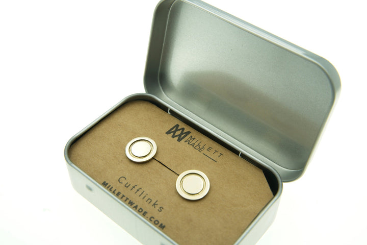 Open metal tin box containing a pair of Round Cufflinks on a cardboard backing labeled "Millett Wade." The inside of the lid features the "Millett Wade" logo and website URL, "MILLETWADE.COM," also written on the cardboard.