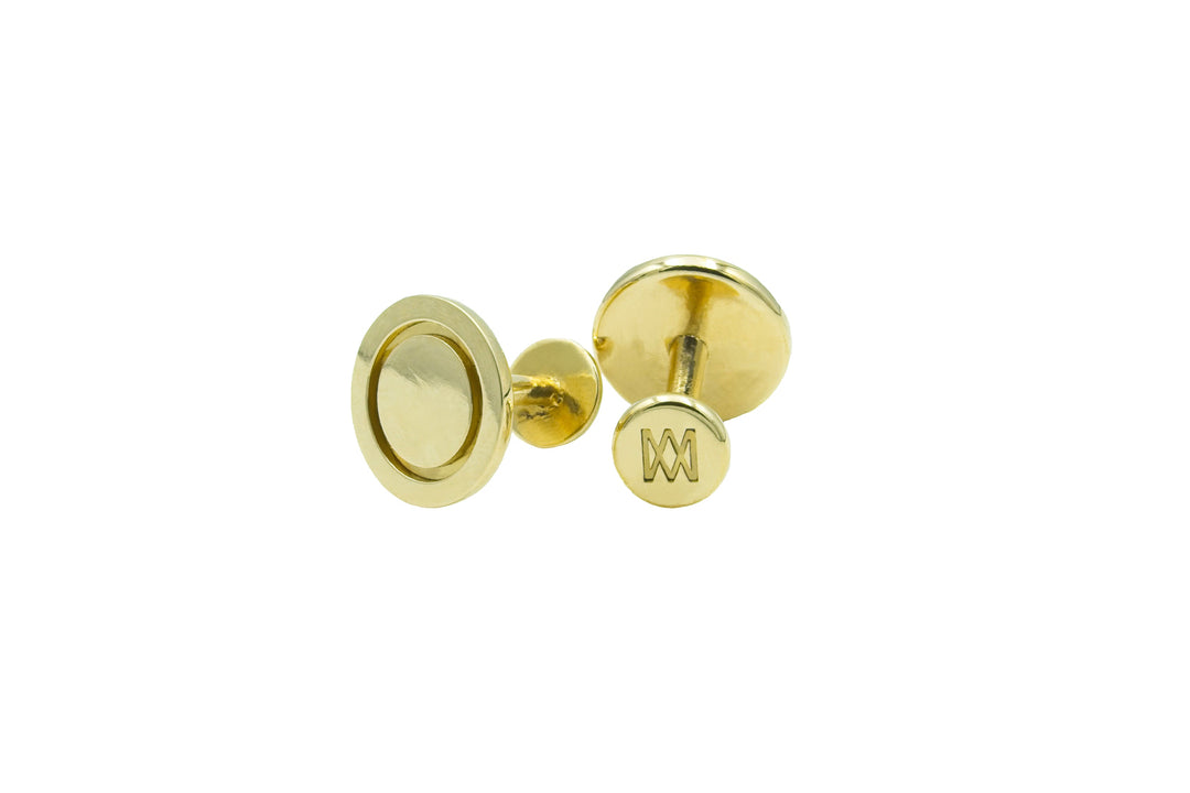 A pair of hand-finished, gold Round Cufflinks crafted from solid brass with a smooth, round design by Millett Wade. One cufflink faces forward, showcasing a circular pattern, while the other reveals its back with an engraved letter "M". Both cufflinks boast a shiny and polished finish.