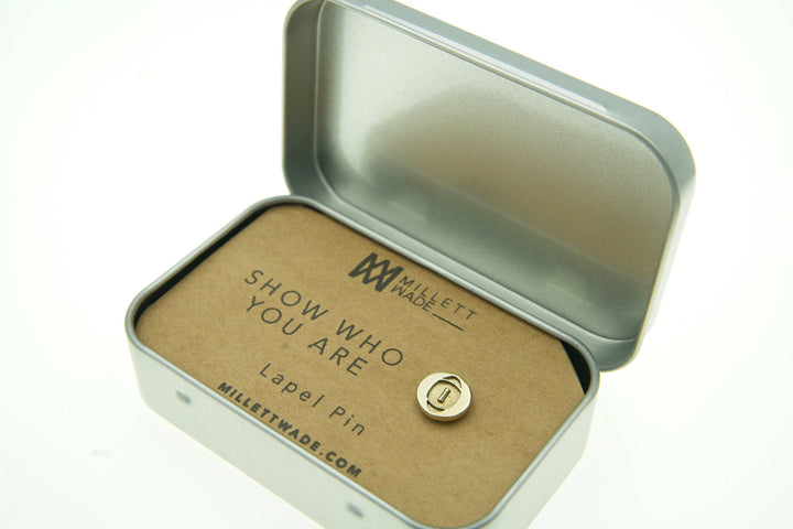 An open metal box showcases a custom pin on a cardboard backing that reads "SHOW WHO YOU ARE" with "Rugby Lapel Pin" below. The box lid is lifted, revealing the branding "Millett Wade" at the top. The website milletwade.com is noted at the bottom. This hand-finished brass lapel pin exudes elegance and identity.