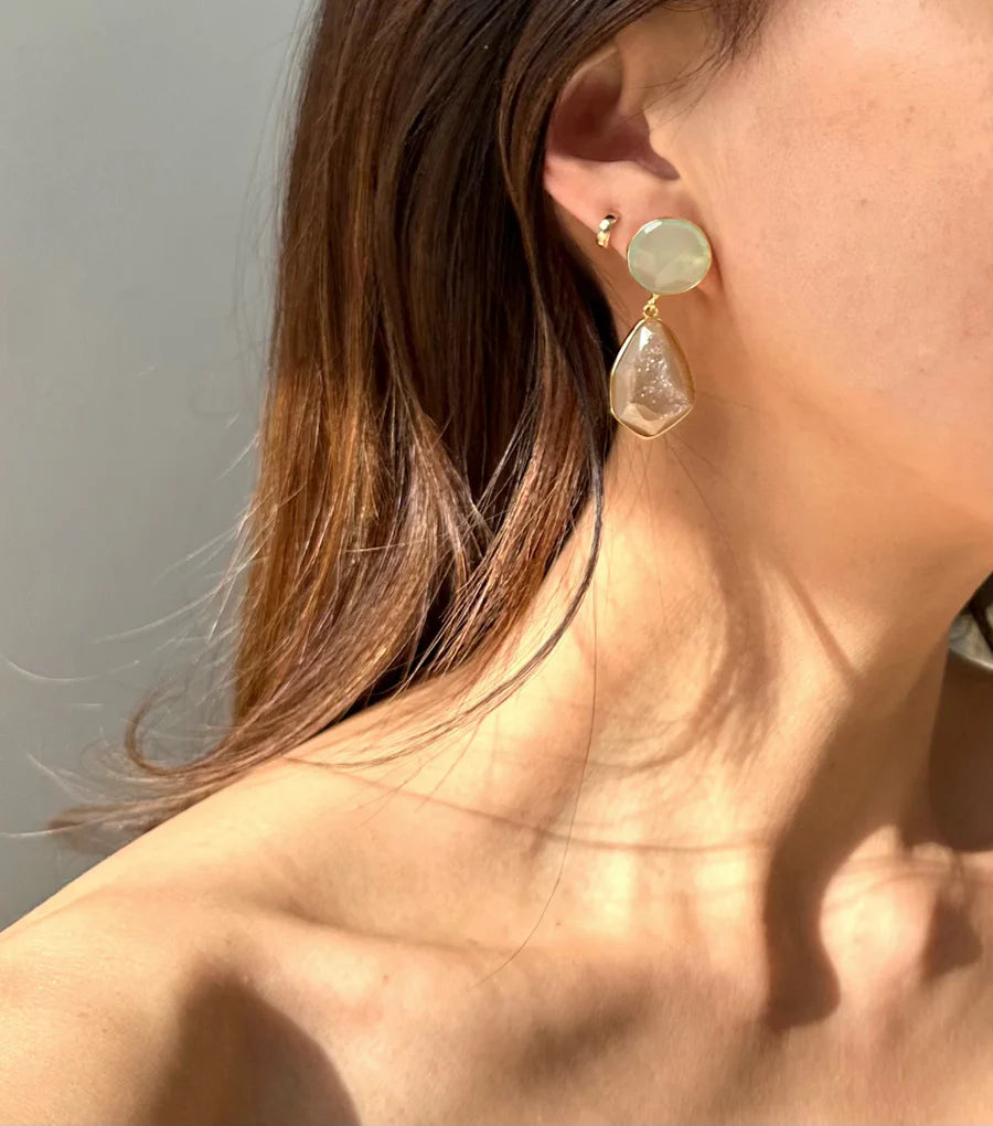 Close-up of a woman wearing Sahara Earrings by Aria V – a round druzy agate gemstone at the earlobe and a teardrop-shaped one hanging below. She has long brown hair, and a second small stud earring in the cartilage of her left ear is also visible.