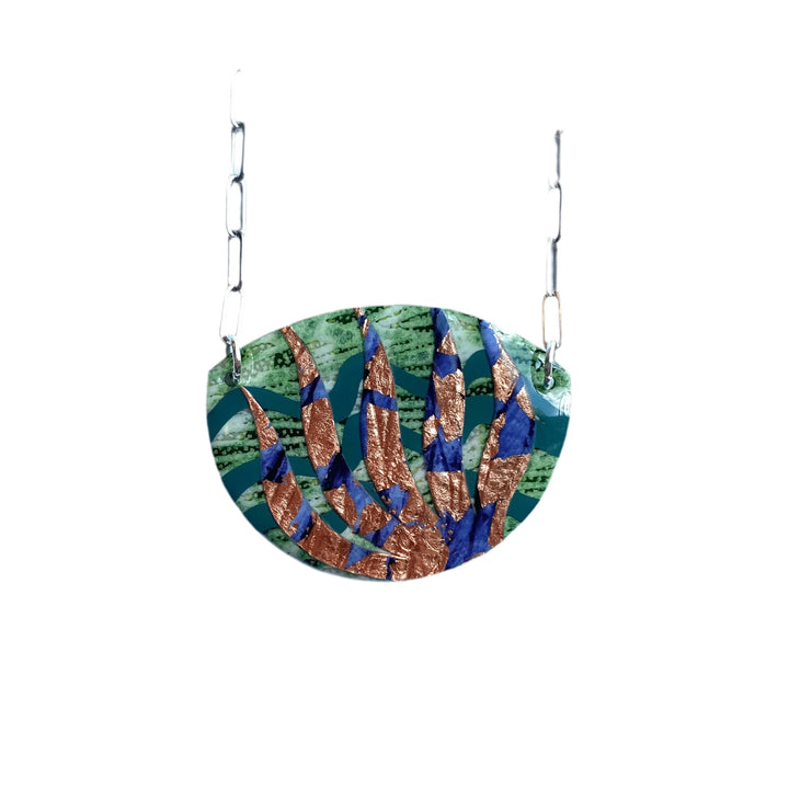 The Rothlú Sáile Batik Textile Necklace in Lime/Indigo/Rose-Gold features a handmade half-circle pendant with a mosaic design, suspended from a silver chain, offering an eco-friendly and distinctive jewelry piece.