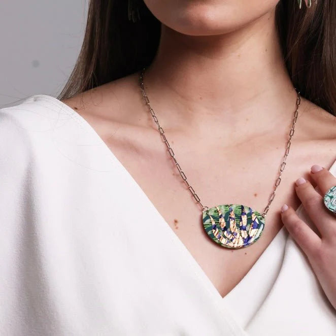 A woman in a white top models Rothlú's Sáile Batik Textile Necklace in Lime/Indigo/Rose-Gold, featuring a large oval pendant with an abstract pattern and a matching ring. Only her lower face and upper torso are visible.