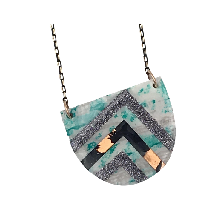 A Rothlú Sayre Batik Textile Necklace in Mint/Black/Copper/Grey Shimmer featuring an intricate geometric design. The design includes black, gold, and glittery silver zigzag patterns on a green and white marbled background. The pendant hangs from a delicate black-gold faceted chain adorned with an eco resin finish.