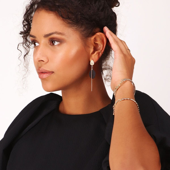 A woman with curly hair in a black top turns to the side, touching her ear to reveal Scribble & Stone's Stem Disc Drop Earrings in 14ct gold-fill. She also wears two gold bracelets and subtle, natural makeup against a plain background.