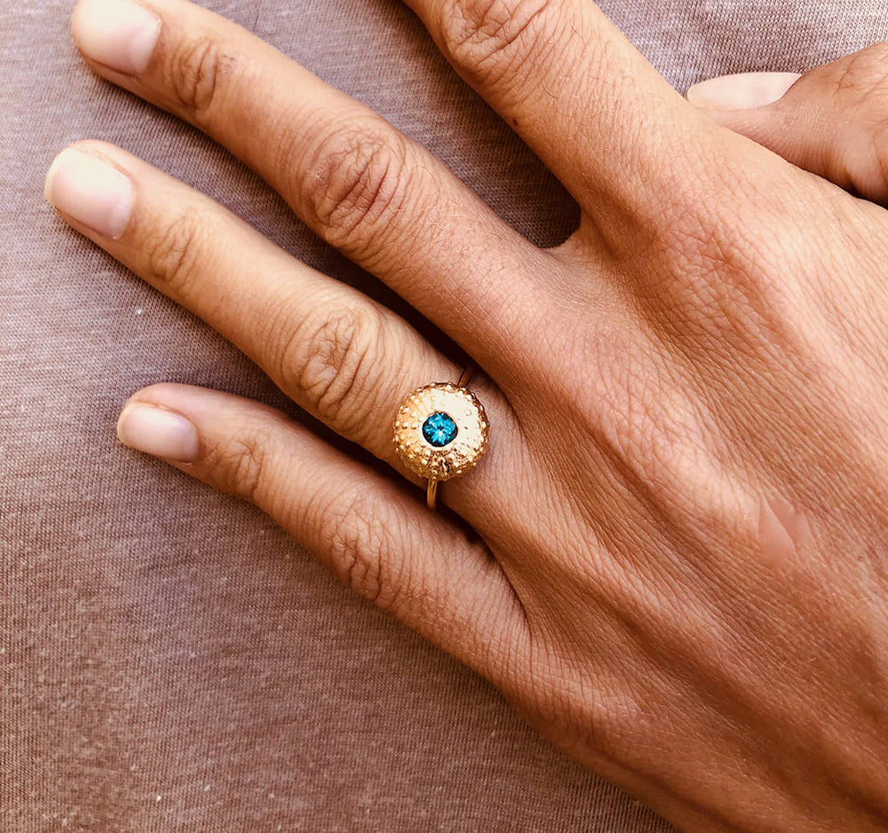 A close-up of a hand on brown fabric, wearing the Jennifer Kinnear Sea Urchin Ring Gold with Stone—a gold plated silver ring featuring a round blue stone surrounded by small clear gems on the ring finger.