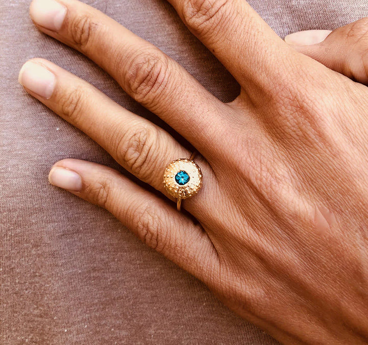 A close-up of a hand on brown fabric, wearing the Jennifer Kinnear Sea Urchin Ring Gold with Stone—a gold plated silver ring featuring a round blue stone surrounded by small clear gems on the ring finger.