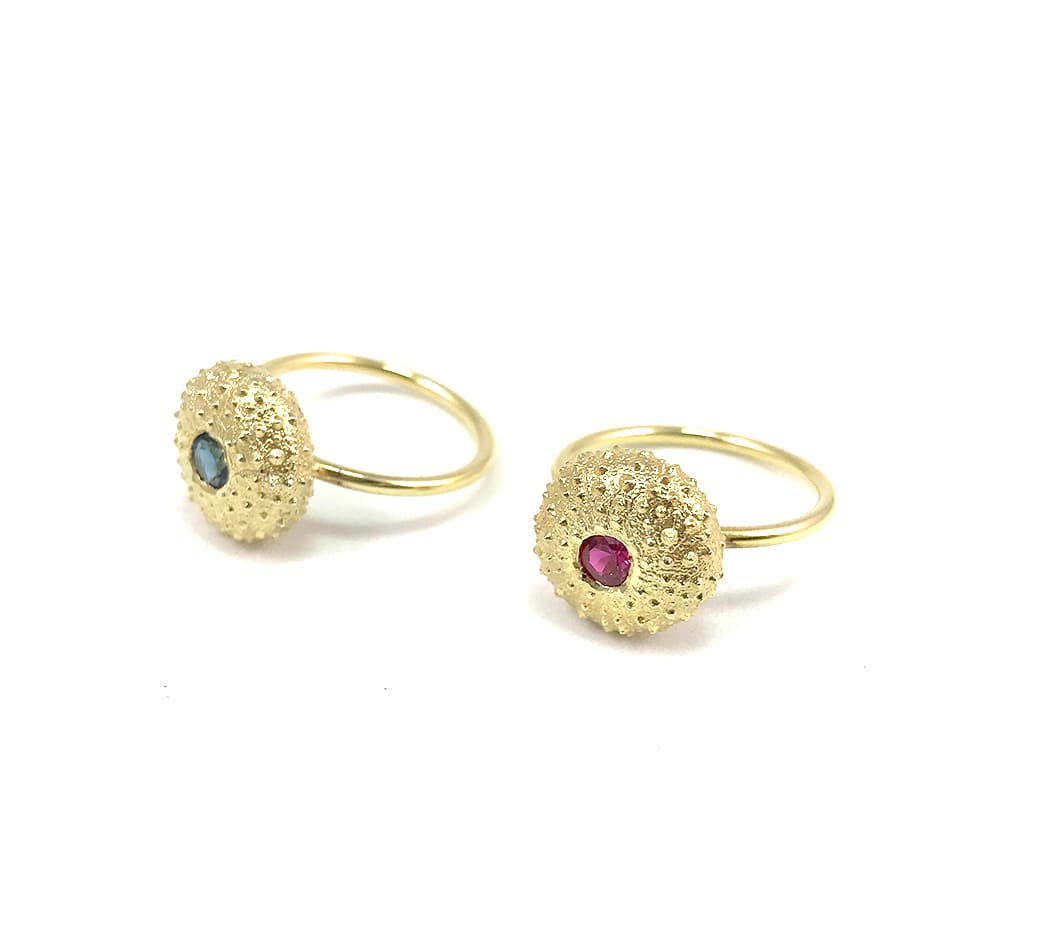 Two Jennifer Kinnear Sea Urchin Ring Gold with Stone rings are shown on a white background, each featuring a textured gold-plated silver circular design with a gemstone—one blue, one pink.