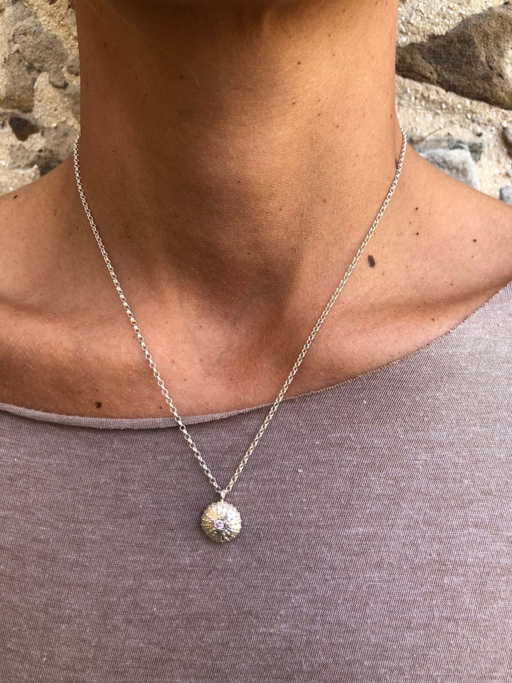 A close-up of a person wearing Jennifer Kinnear's Sea Urchin Necklace with Stone (silver or gold), featuring a round, textured pendant with a gemstone at the center. The person wears a taupe top and stands against a stone wall.
