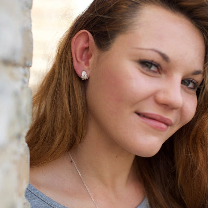 A woman with long brown hair smiles at the camera. She is wearing a gray top, Sea Urchin Wrap Stud Earrings by Jennifer Kinnear, and a delicate necklace. Her head is slightly tilted, and there is a stone wall to her left. The background is softly blurred.