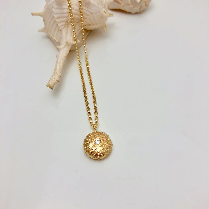 The Jennifer Kinnear Sea Urchin Necklace with Stone, available in silver or gold, features a round, textured sea urchin-inspired pendant with a central gem and is shown next to a light seashell on a white surface.