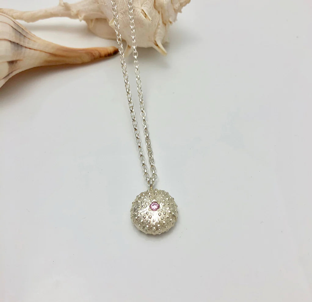 The Jennifer Kinnear Sea Urchin Necklace with Stone, shown in silver and featuring a pink gemstone at its center, is displayed on a white surface beside seashells.