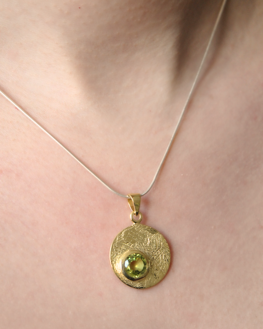 A close-up of a person's neck wearing the Gallardo & Blaine Designs Selene Necklace Gold - Small, featuring a thin chain with a round, textured gold pendant set with a green gemstone.