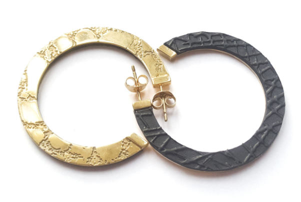 A pair of Sara Gunn Skin Etched Hoop Earrings is shown. Each earring is half gold-plated etched brass and half dark gray, joined together in a circle. The gold sections have an engraved pattern, while the gray sections have a textured pattern. The earrings have butterfly backings.