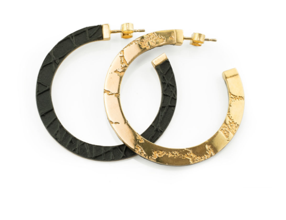 A pair of Skin Etched Hoop Earrings by Sara Gunn is shown on a white background. One hoop features embossed black leather with a textured surface, while the other is crafted from gold-plated etched brass with an irregular design. Both earrings have stud backings.