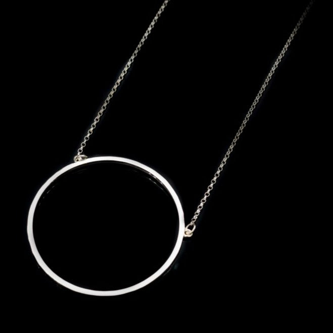 A delicate Lynsey de Burca Ancaire Silver Neckpiece features a thin, hand-forged circular pendant, suspended by a fine chain. The image is set against a black background, emphasizing the minimalist and elegant design of this jewelry piece.