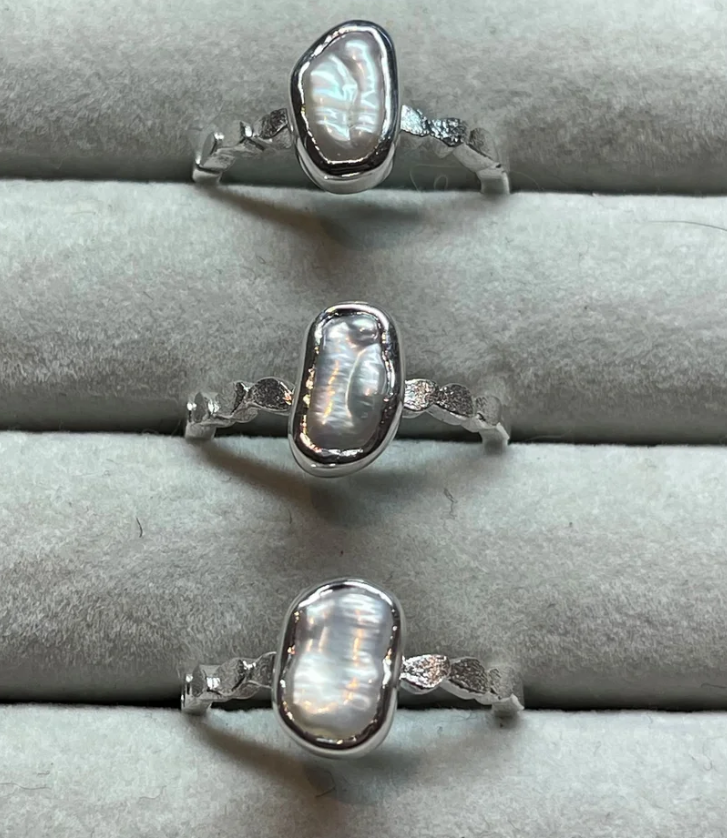 Three silver rings with polished irregular oval baroque keshi centre stones displayed on a grey ring stand.