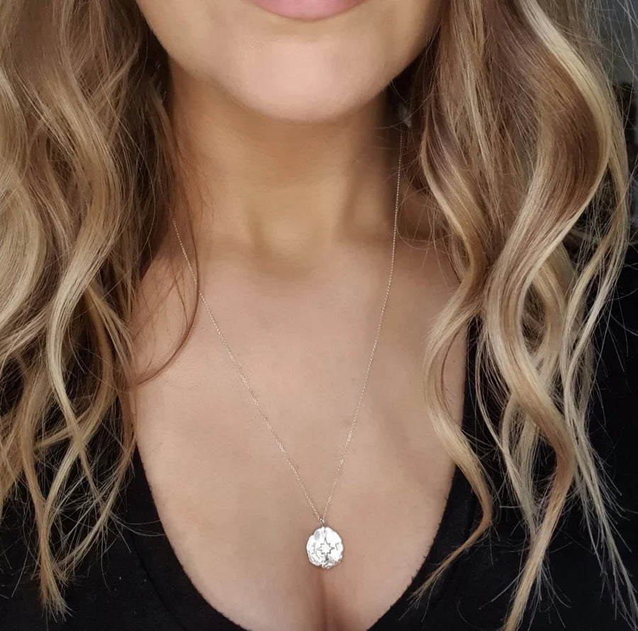 A woman with wavy blonde hair wears the Pale Raven Compass Coin Silver Necklace with a black V-neck top, highlighting her neck, collarbone, and part of her chest.