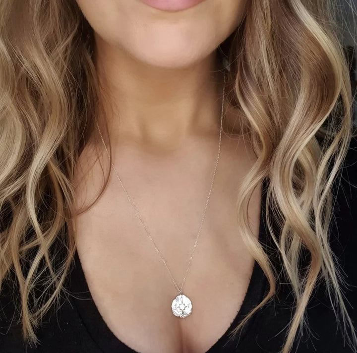 A woman with wavy blonde hair wears the Pale Raven Compass Coin Silver Necklace with a black V-neck top, highlighting her neck, collarbone, and part of her chest.