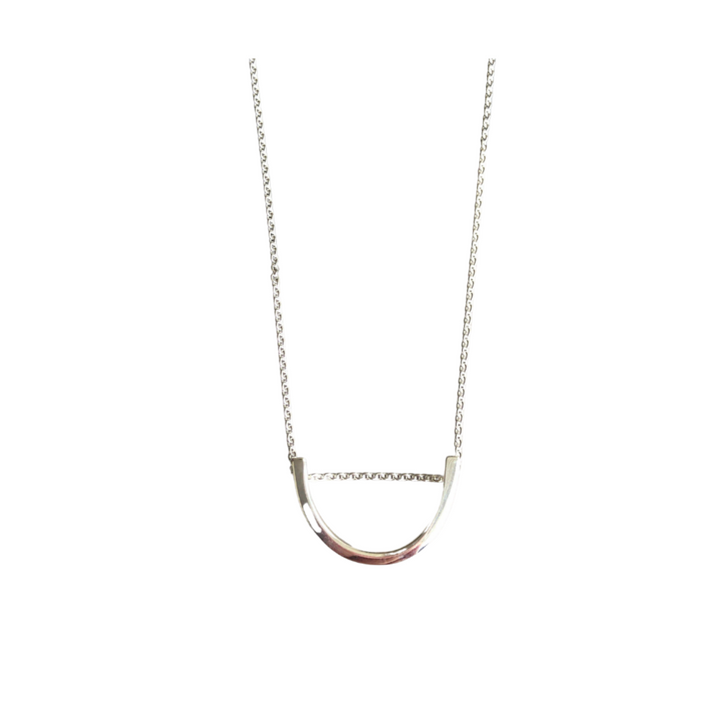 The Angela O'Keefe Silver Curve Pendant features a U-shaped pendant. Handmade in Ireland, the symmetrical and smooth pendant connects to a delicate sterling silver chain on both ends. The design is simple and elegant, emphasizing clean lines and a modern aesthetic against a plain white background.