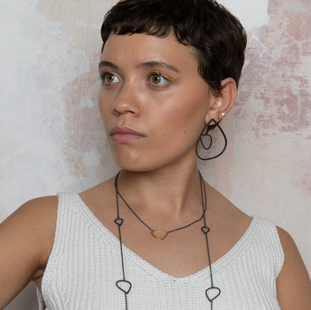 A person with short dark hair wears a white knit tank top, large geometric earrings, and a matching Gold Wing Pendant with Oxidised Silver Chain by Leoma Drew. They stand against a lightly textured wall and gaze to the left with a neutral expression.