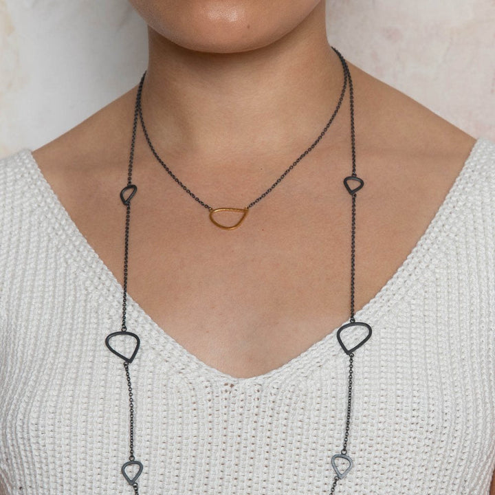 A person wearing a white knit top is shown from the shoulders down, accessorized with two necklaces. One shorter necklace features Leoma Drew's Gold Wing Pendant on an Oxidised Silver Chain, while the longer necklace showcases large, irregularly shaped links spaced along the chain. The background is light-colored.