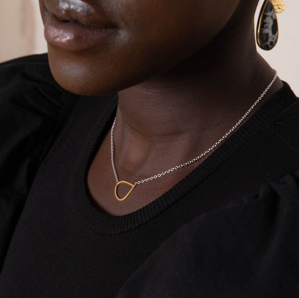 A close-up image of a person wearing a black top, showcasing a Leoma Drew Gold Wing Pendant with Silver Chain. The focus also captures a portion of yellow drop earrings with black accents. The background is blurred.