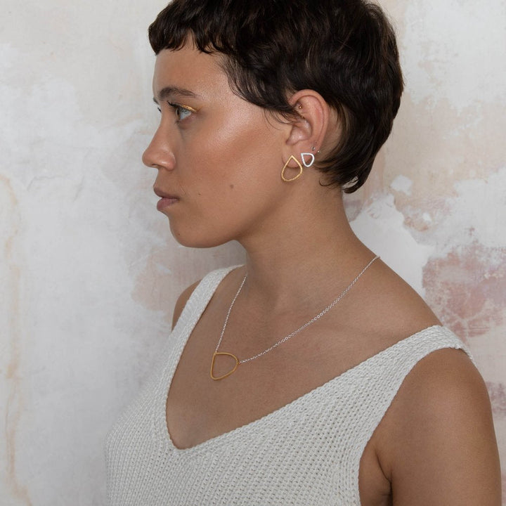 A person with short dark hair is wearing a white knit tank top, gold earrings in the shape of abstract teardrops, and a matching Leoma Drew Gold Wing Pendant with Silver Chain. They are facing left against a textured beige and white background.