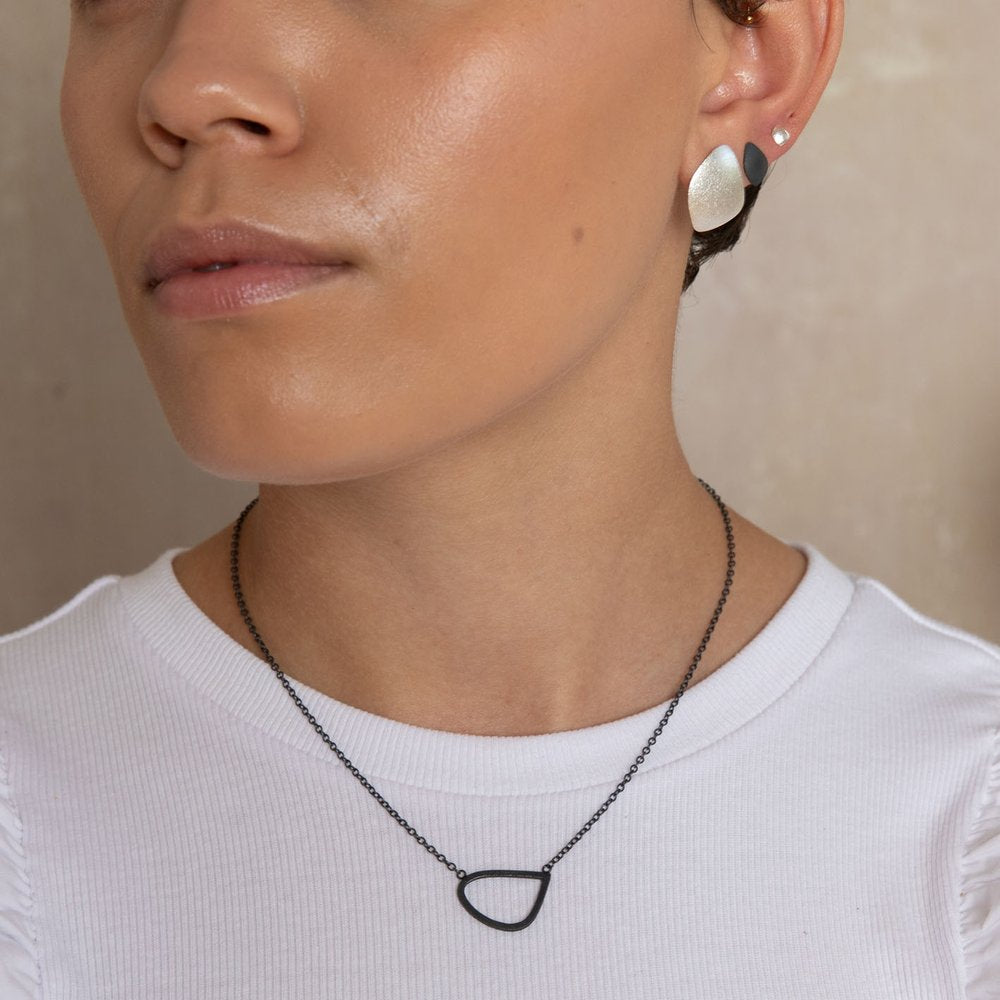 Close-up of a person wearing a white top, featuring minimalist earrings with metallic finishes and a black chain necklace with a hand-crafted pendant shaped like a rounded triangle. The background is neutral, and the person’s face is partially visible. They are wearing the Single Wing Pendant Oxidised Silver by Leoma Drew.