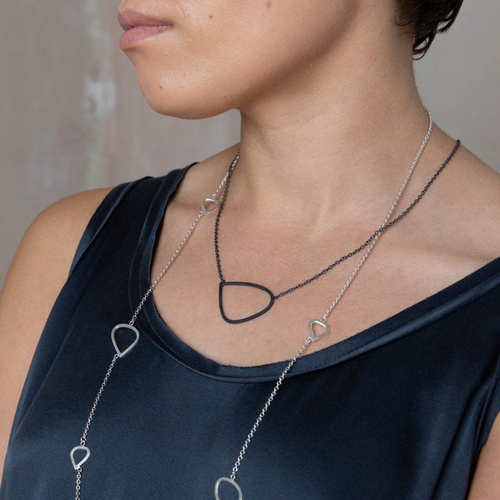 A person wearing a dark sleeveless top with layered necklaces. One necklace is a handcrafted black piece with a geometric pendant, and the other is a Single Wing Pendant Oxidised Silver by Leoma Drew, adorned with several similar geometric shapes. The background is an out-of-focus neutral tone.