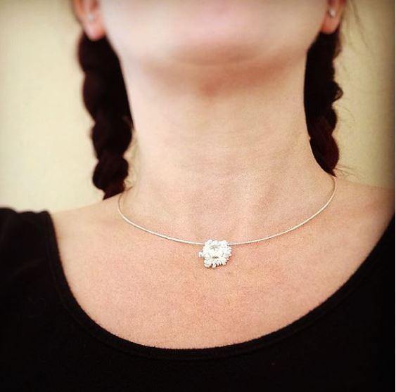 A close-up of a woman wearing an Abbie Dixon Spore Small Pendant Black & Gold. She has two braids, and her top is black with a wide neckline. The background is plain, allowing the focus to be on the pendant.