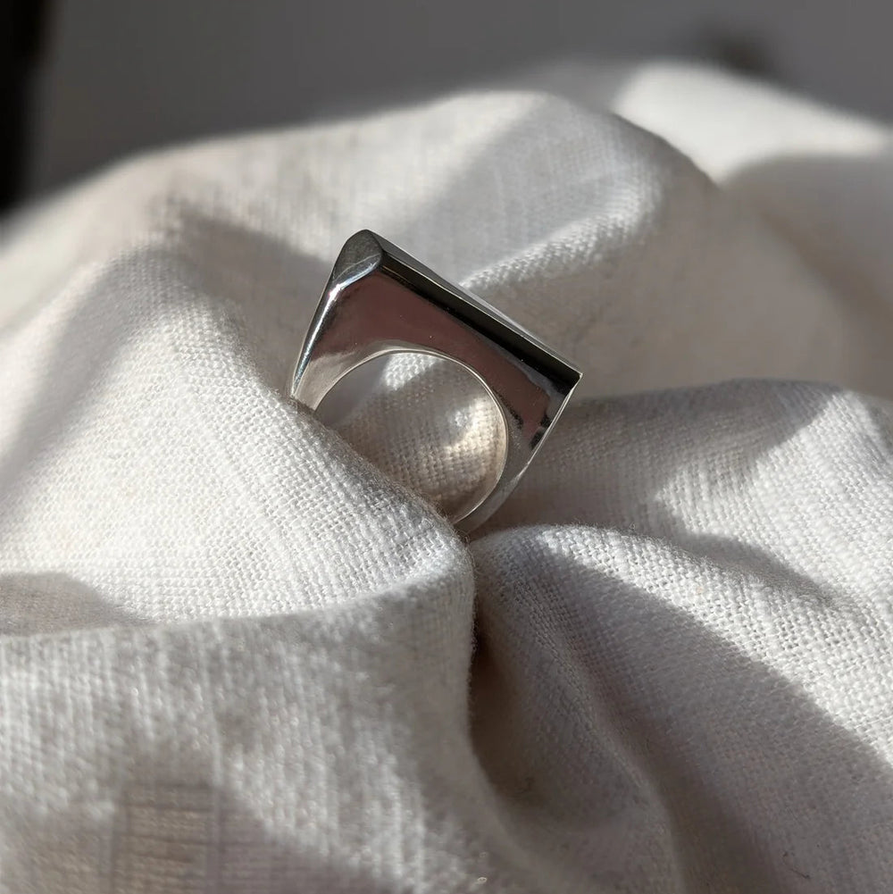 The Smooth Lines Chunky Ring by Becca Calmont, crafted from recycled sterling silver with a flat rectangular top and bold geometric shape, sits on soft white textured fabric bathed in natural light.