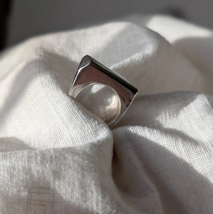 The Smooth Lines Chunky Ring by Becca Calmont, crafted from recycled sterling silver with a flat rectangular top and bold geometric shape, sits on soft white textured fabric bathed in natural light.