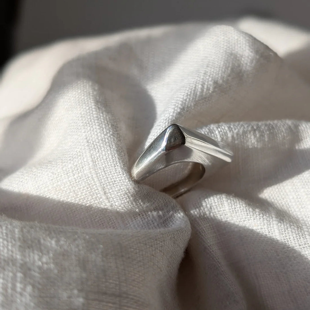 The Smooth Lines Chunky Ring by Becca Calmont, crafted from recycled sterling silver, sits on a soft off-white fabric, illuminated by sunlight that casts gentle shadows and highlights across its textured surface.