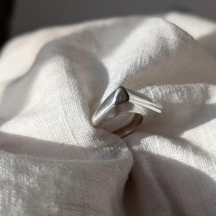 The Smooth Lines Chunky Ring by Becca Calmont, crafted from recycled sterling silver, sits on a soft off-white fabric, illuminated by sunlight that casts gentle shadows and highlights across its textured surface.