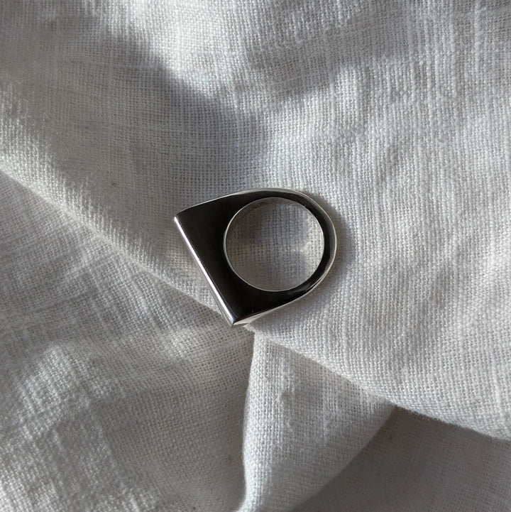The Smooth Lines Chunky Ring by Becca Calmont, made from recycled sterling silver with a flat top and bold geometric design, sits on white textured fabric glowing in natural light—a true statement piece.