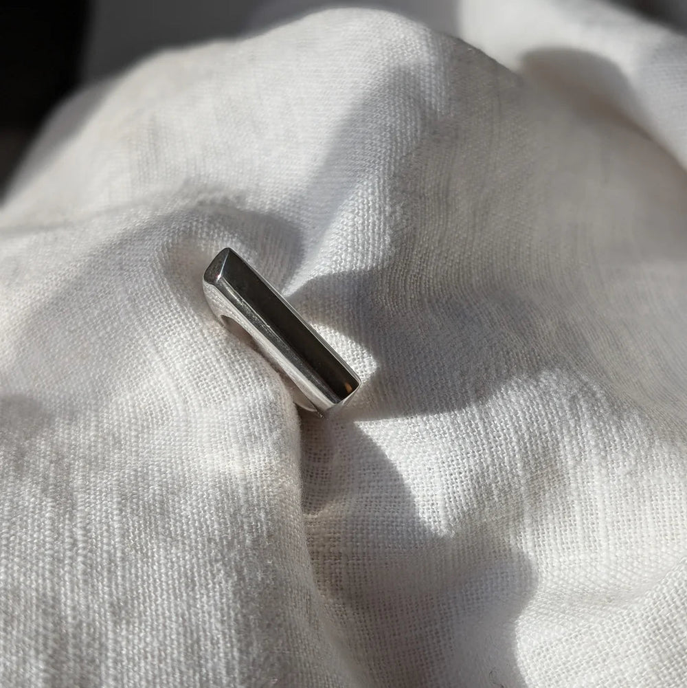 The Smooth Lines Chunky Ring by Becca Calmont, made from recycled sterling silver, stands upright on softly wrinkled white fabric, illuminated by natural light and casting gentle shadows.