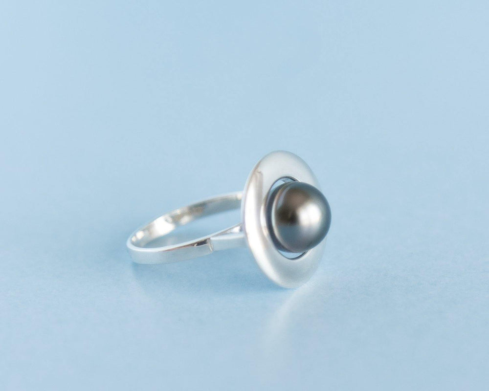 A custom-made sterling silver ring with a unique design featuring a Tahitian pearl set in the center of a flat, circular disc. The Home Planet Silver Tahitian Pearl by Cosmic Boulevard is placed on a light blue surface, highlighting its sleek and modern appearance.