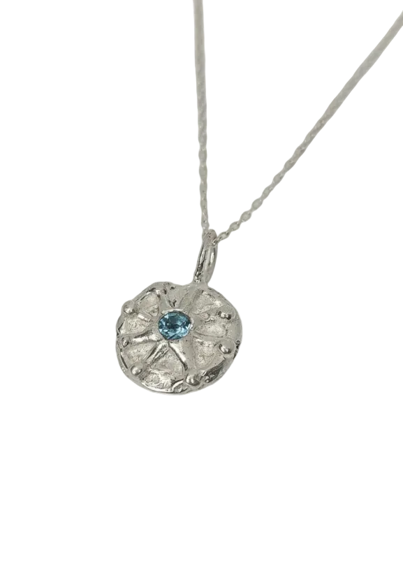 A silver pendant necklace featuring a textured, starburst-like silver medallion with a small round light blue gemstone in the center, suspended from a delicate silver chain against a plain white background.